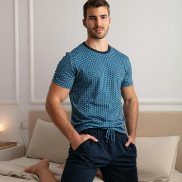 Man wearing a blue patterned shirt and dark shorts sitting on a bed.